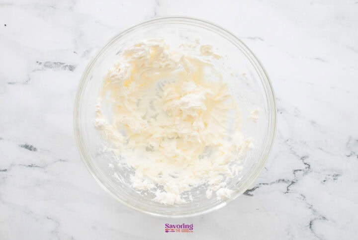 A glass bowl on a marble surface contains the beginnings of chocolate lasagna, with partially mixed cream cheese and butter suggesting layers of indulgence.