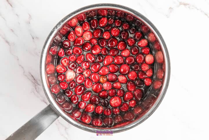 A saucepan filled with sugared cranberries in sugar water rests on a white marble surface.