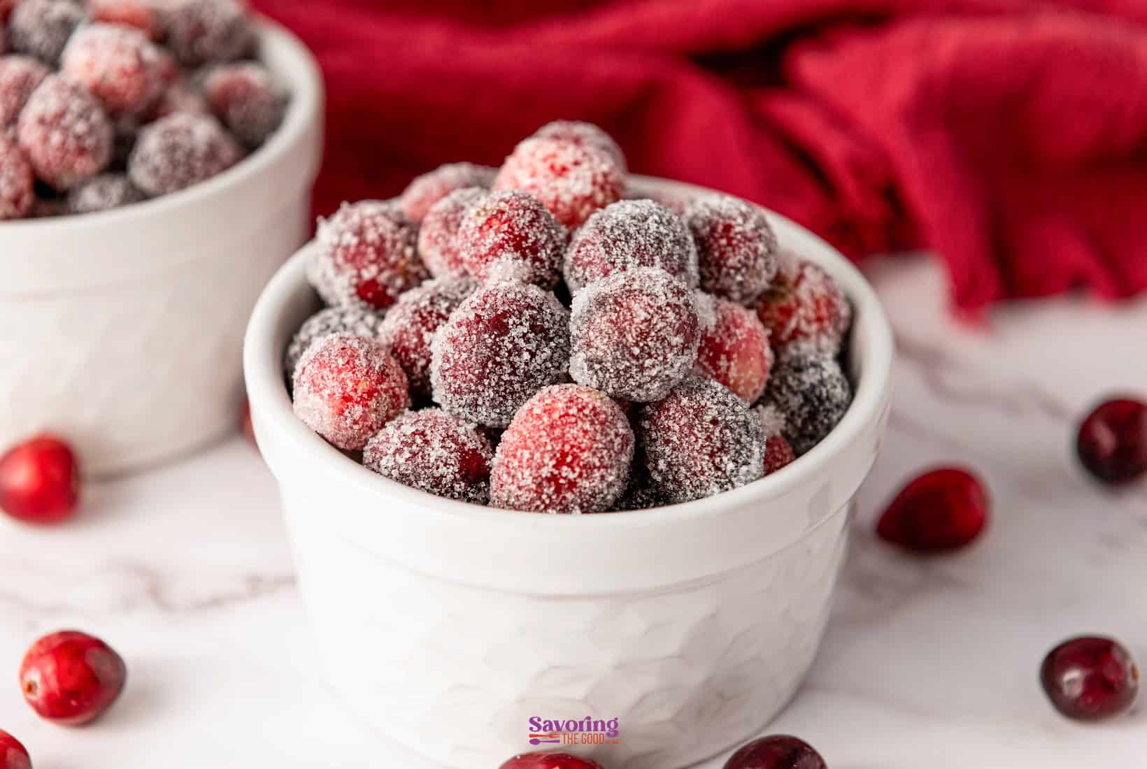 A white bowl brimming with glistening sugared cranberries sits on a marble surface, while a few escapees dot the scene. A vibrant red cloth in the background adds a festive touch to this delightful display.