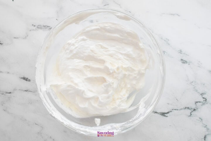 A glass bowl filled with whipped cream sits enticingly on a marble countertop, hinting at the indulgent layers of chocolate lasagna to come.