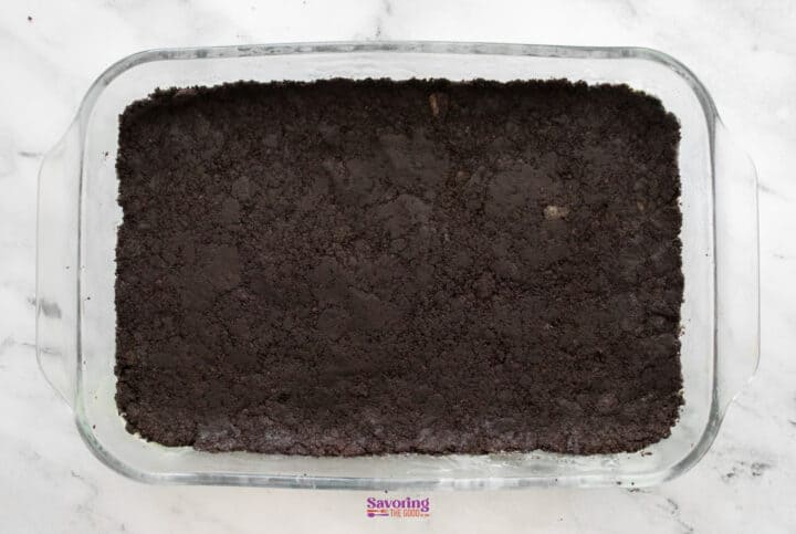 A glass baking dish filled with a layer of crumbled chocolate cookie crust, reminiscent of a decadent chocolate lasagna, rests on a marble countertop.