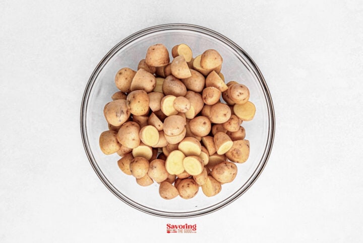 A clear glass bowl filled with perfectly roasted baby potatoes, sliced in half, rests on a pristine white surface.