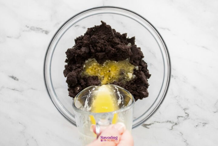 A glass bowl contains dark cookie crumbs being mixed with melted butter, forming the perfect base for a rich chocolate lasagna.