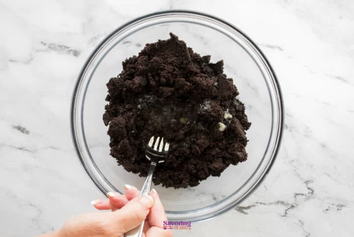 A hand holding a fork is mixing crushed cookies and butter for a delectable Chocolate Lasagna in a clear glass bowl on a marble surface.