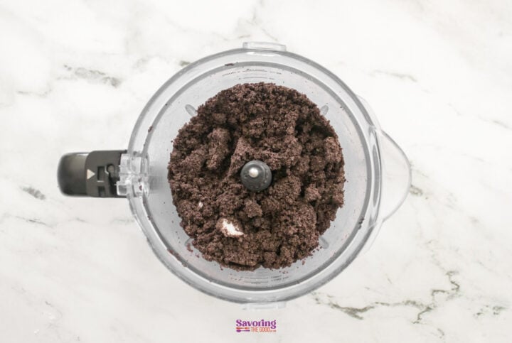 A food processor brimming with ground chocolate cookies sits on a marble countertop, ready to create the base for a decadent chocolate lasagna.