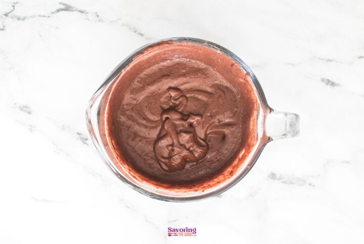 A glass measuring cup filled with rich chocolate cake batter, reminiscent of decadent layers in a chocolate lasagna, sits on a pristine white marble surface.