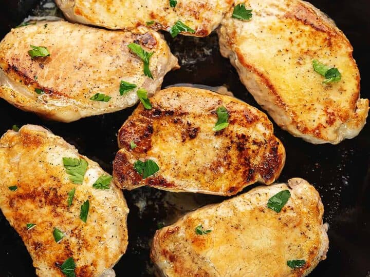 A black skillet with six golden-brown, cooked pork chops, garnished with fresh herbs.
