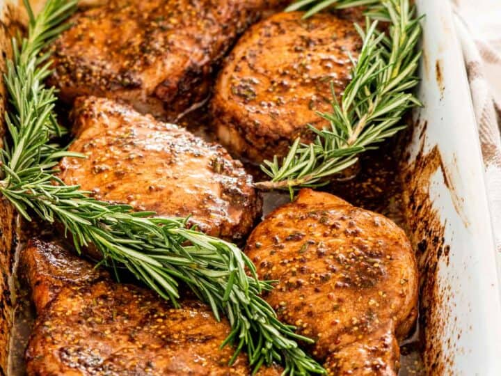 Baked pork chops seasoned with herbs and spices, garnished with fresh rosemary, in a white baking dish.