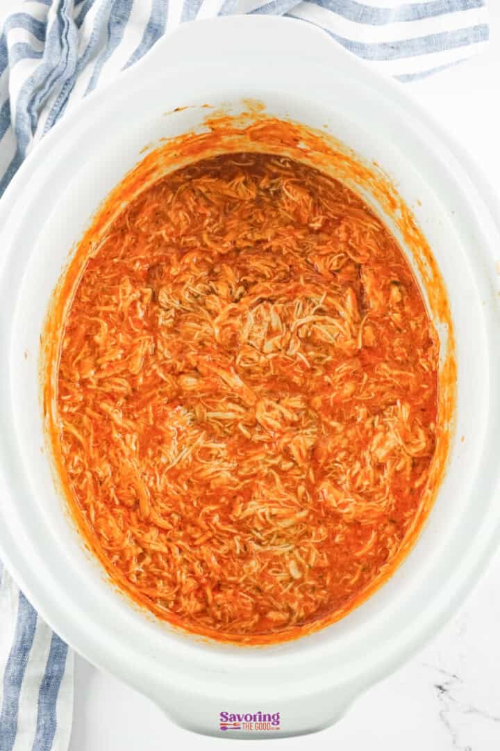 A tantalizing slow cooker Buffalo chicken dip recipe awaits, with shredded chicken basking in a rich red sauce, all resting invitingly on a striped cloth.