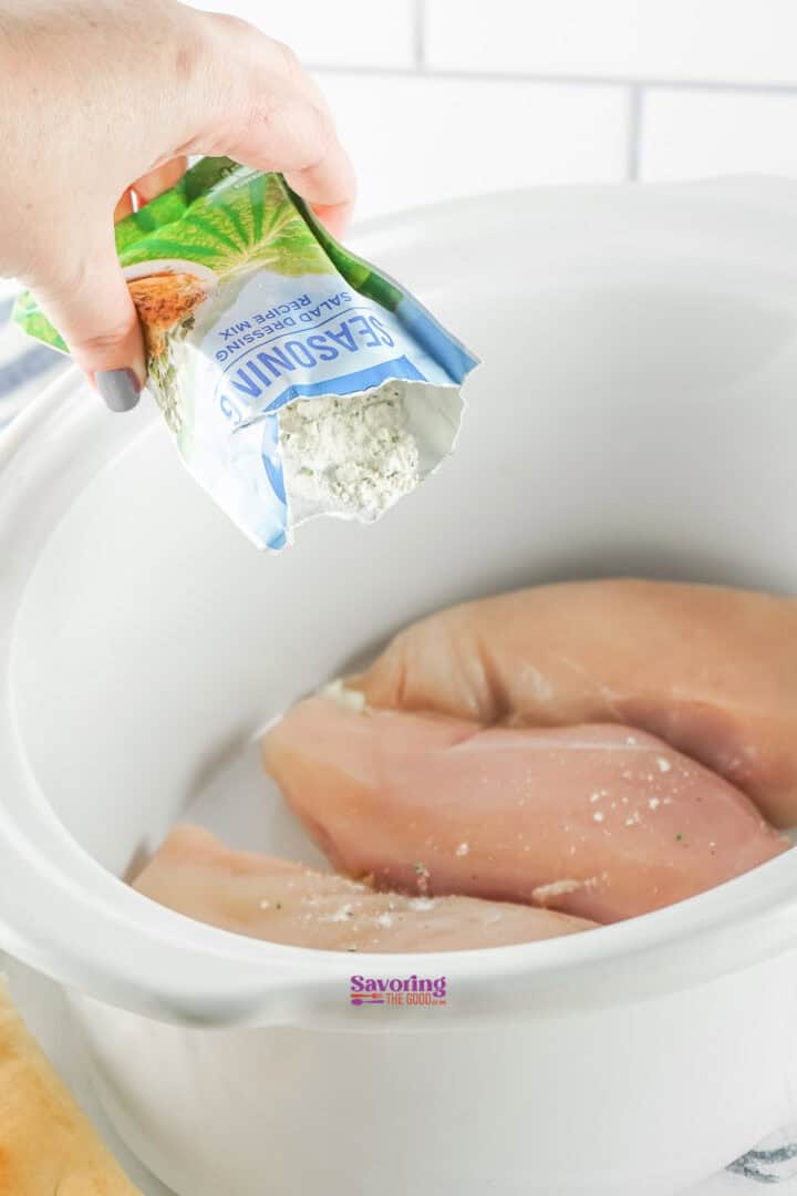 A hand pours a Buffalo-flavored seasoning mix over raw chicken breasts in a white slow cooker, setting the stage for a delicious Slow Cooker Buffalo Chicken Dip recipe.