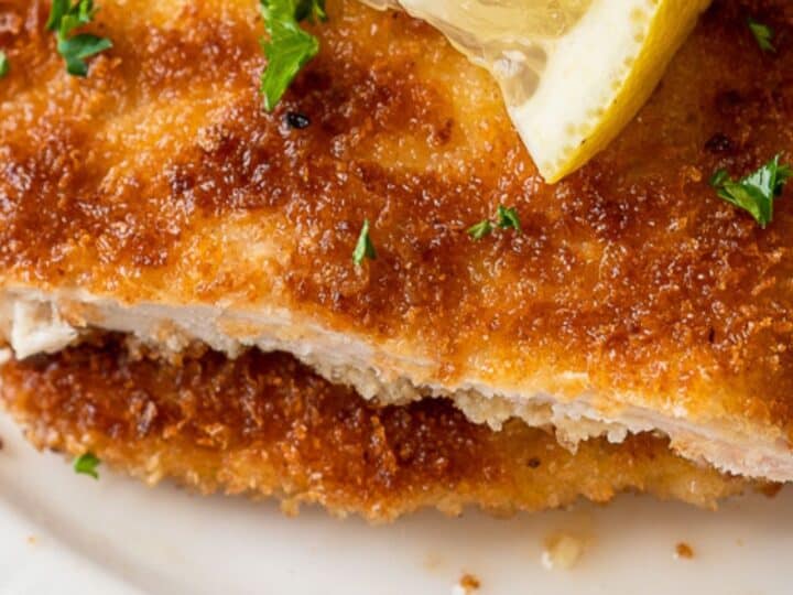 Close-up of a breaded and pan-fried meat slice topped with lemon and parsley on a white plate.