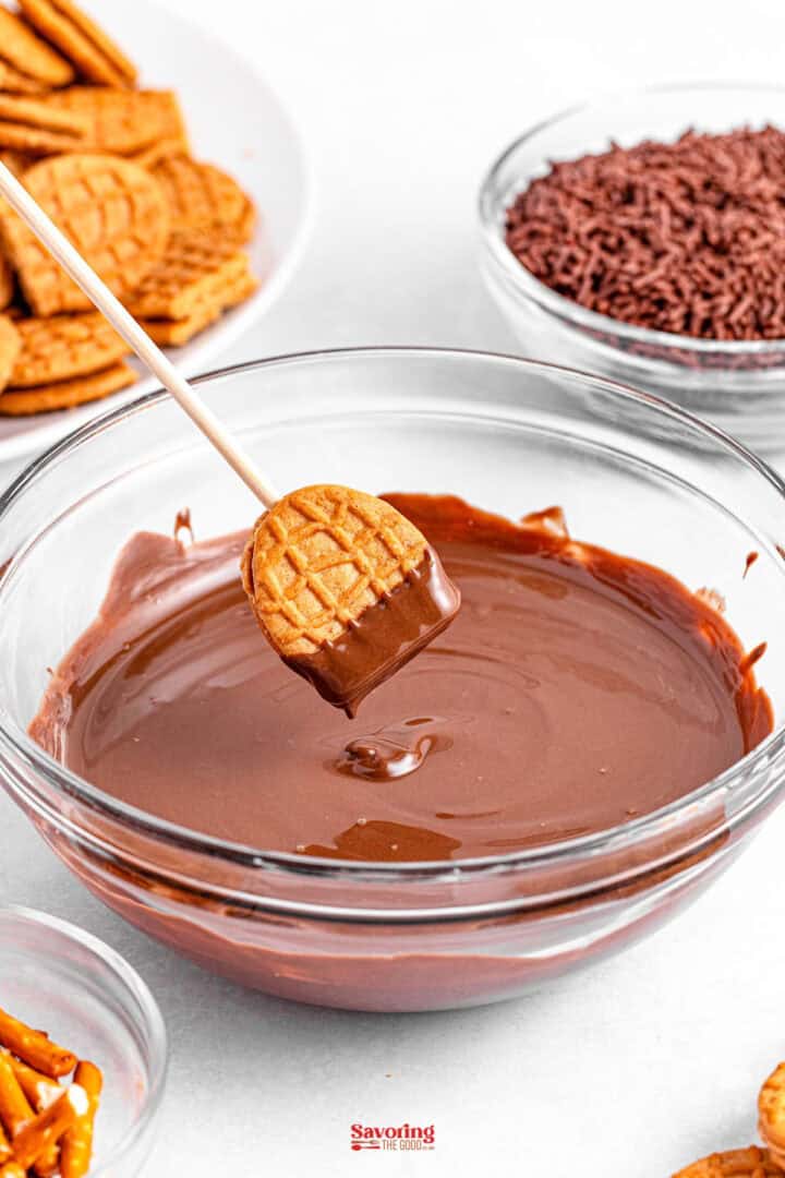 An acorn cookie on a stick is dipped into a bowl of melted chocolate, surrounded by pretzels and chocolate sprinkles.