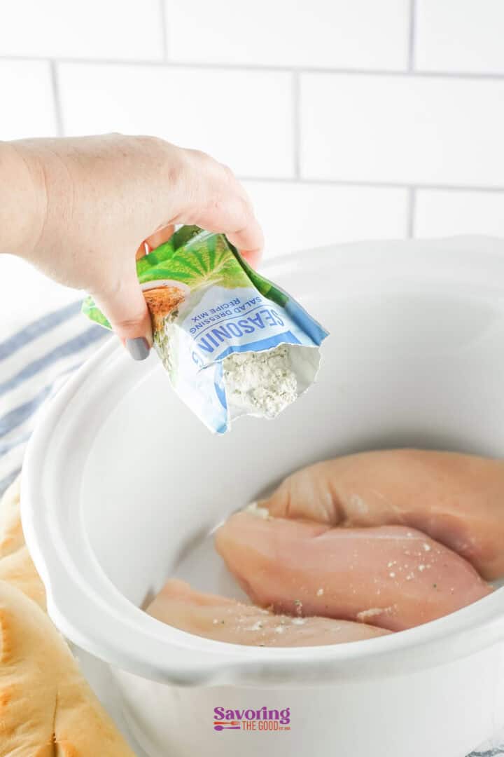 A hand pours seasoning from a packet onto raw chicken breasts in a slow cooker.