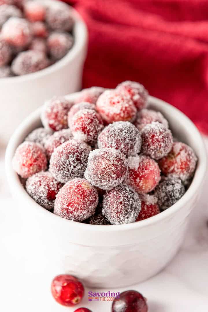 A white bowl brimming with glistening sugared cranberries rests against a vibrant red background, capturing a festive allure.
