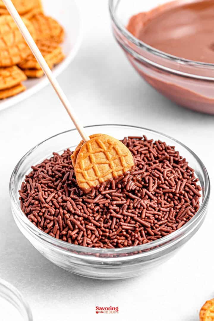 A small acorn cookie on a stick is being dipped into a bowl of chocolate sprinkles, with another bowl of melted chocolate nearby.
