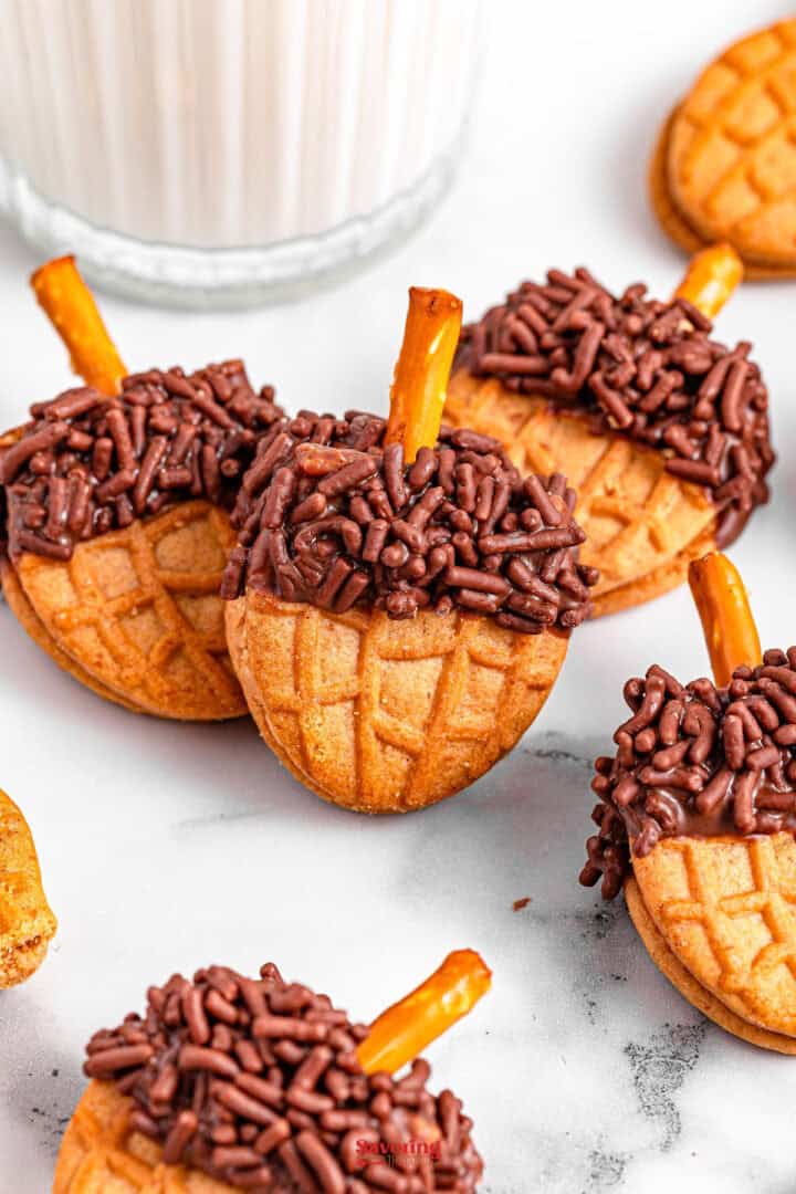 Acorn cookies adorned with chocolate sprinkles and pretzel stick stems rest on a marble surface, perfectly complemented by a glass of milk in the background.