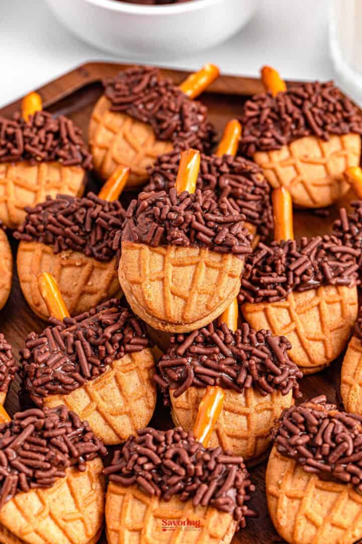 Acorn cookies adorned with chocolate sprinkles and pretzel sticks are artfully arranged on a wooden tray.