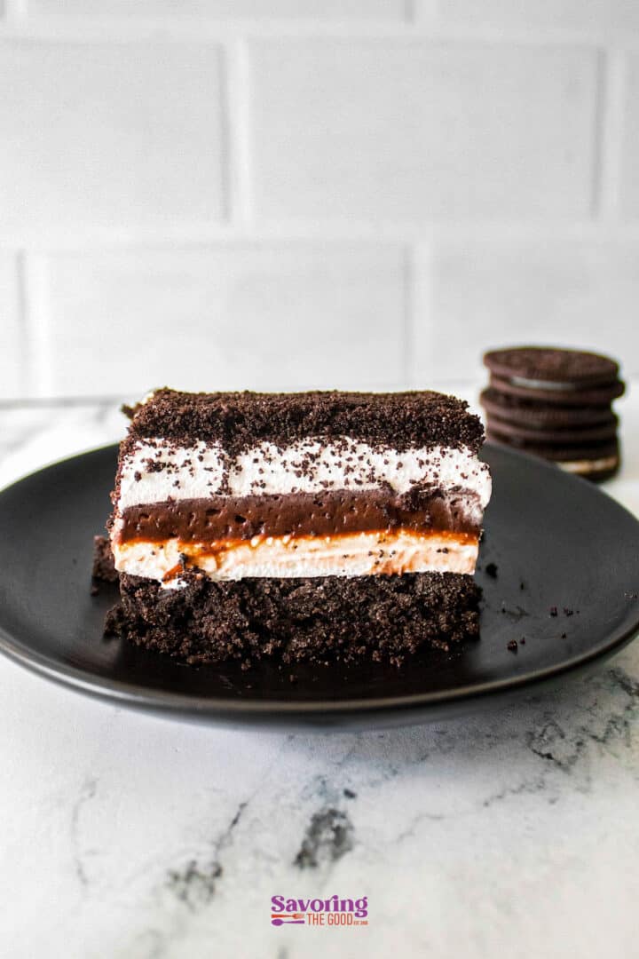 Slice of layered chocolate lasagna dessert with Oreo crumbs, whipped cream, chocolate, and cream cheese on a black plate; Oreos in the background.