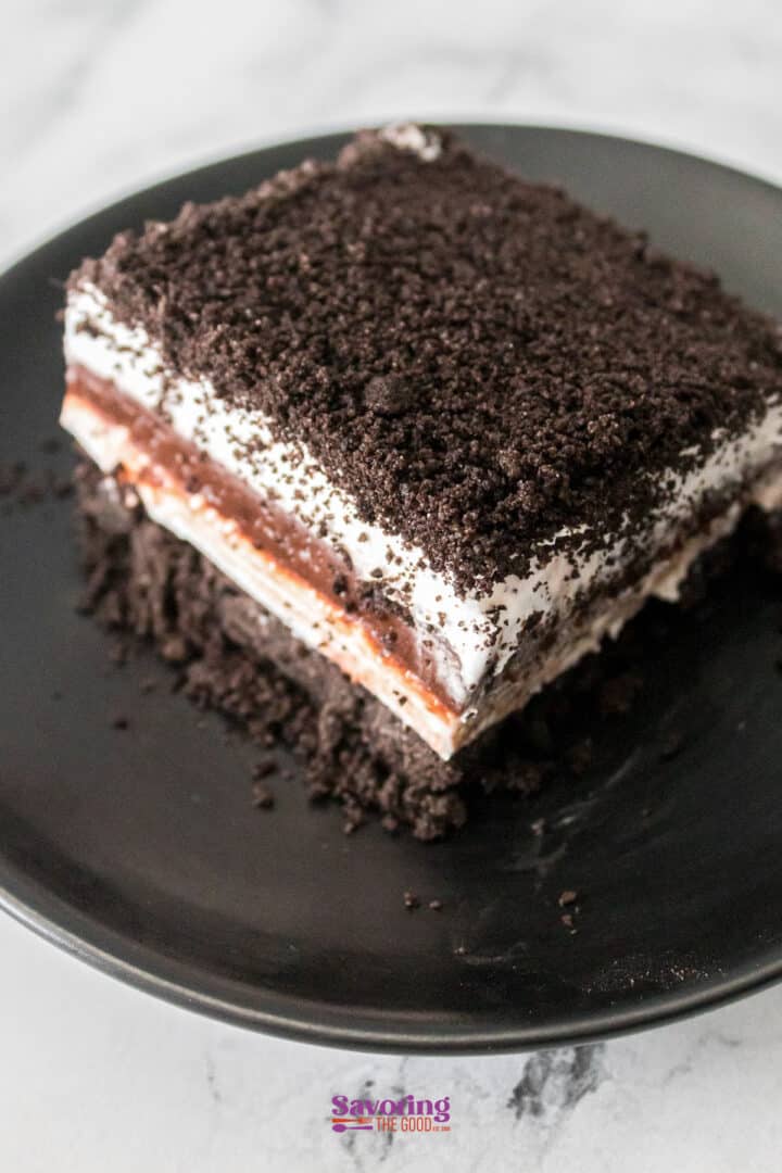A slice of chocolate lasagna with layers of crushed cookies, a creamy filling, and decadent chocolate rests elegantly on a black plate.