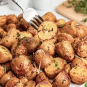 A fork holds a slice of seasoned roasted baby potato over a pile of roasted potato chunks garnished with fresh herbs and melted cheese.