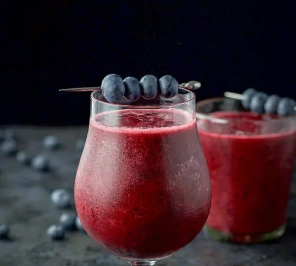 Two glasses of blueberry smoothie garnished with skewered blueberries, set against a dark background.