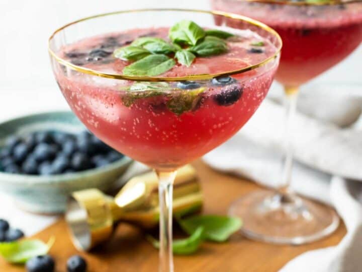 Two glass goblets filled with a pink berry cocktail, garnished with fresh basil leaves. Blueberries scattered around on a wooden surface.