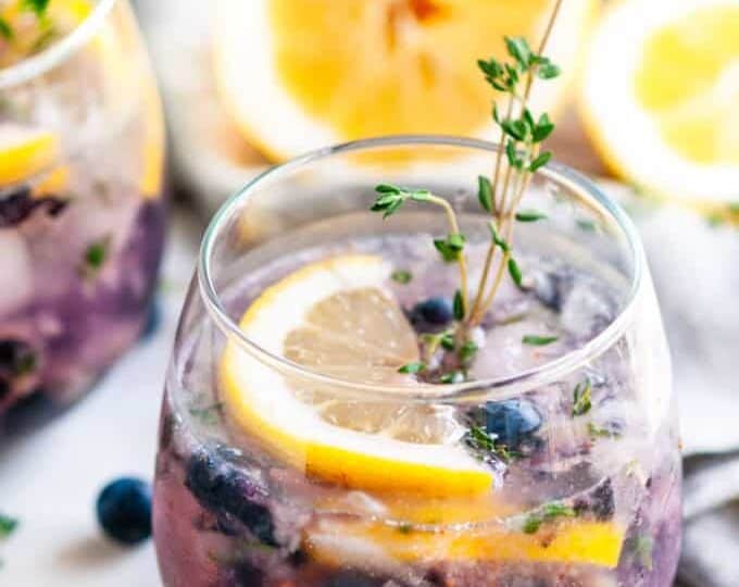 A glass of blueberry lemonade garnished with lemon slices and a sprig of thyme, with another glass and a sliced lemon in the background.
