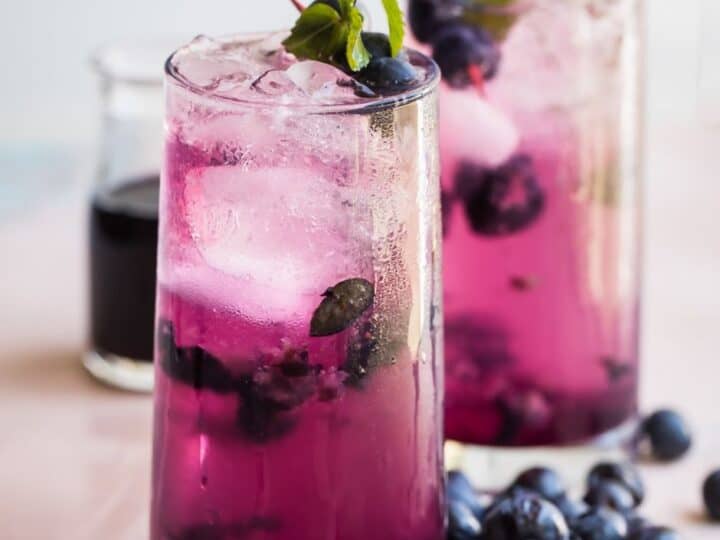 Two glasses of blueberry lemonade garnished with mint leaves and ice, surrounded by fresh blueberries.