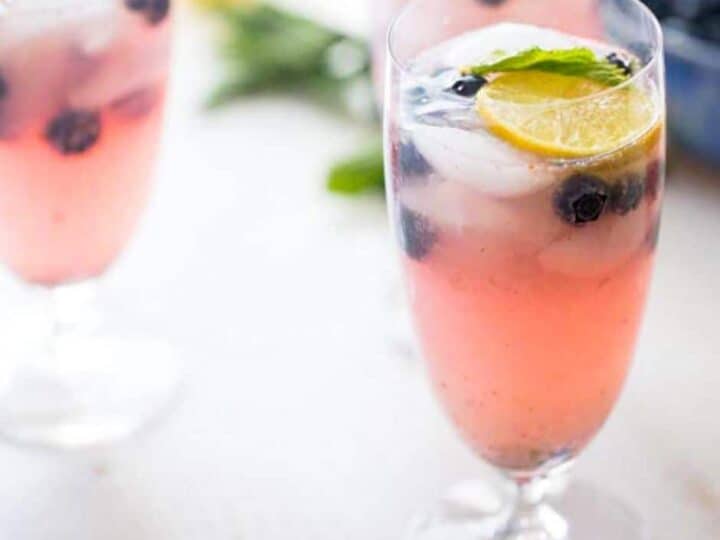 A glass of pink lemonade with ice, blueberries, a lemon slice, and a mint leaf. Other glasses are blurred in the background.