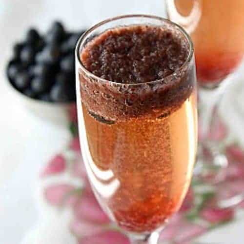A glass of red grape slushie served in a champagne flute, with blueberries in a bowl in the background.