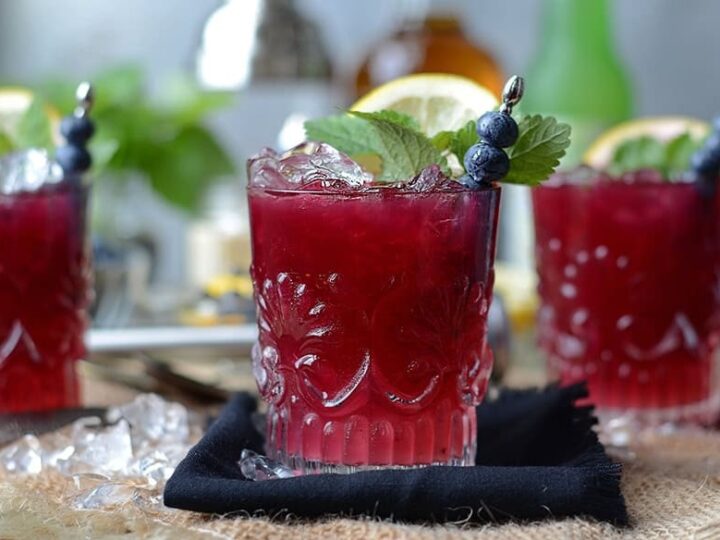 A glass of red cocktail with ice, garnished with blueberries, mint, and a lemon slice. Two similar drinks are in the background.