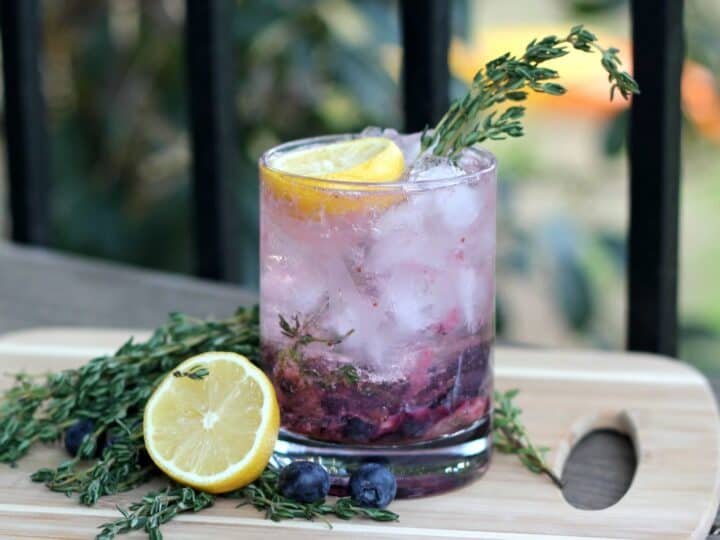 A cocktail with ice, lemon slices, blueberries, and thyme garnish, on a wooden board with additional thyme, lemon, and blueberries.