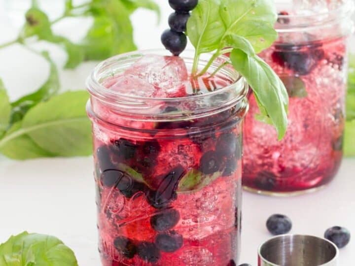 Two mason jar drinks with ice, blueberries, and basil garnishes. A metal jigger and blueberries are nearby on a white surface.