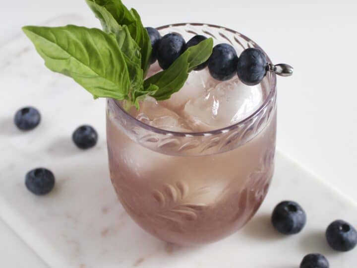A glass of iced blueberry drink garnished with basil leaves and a skewer of blueberries sits on a marble tray, surrounded by scattered blueberries.