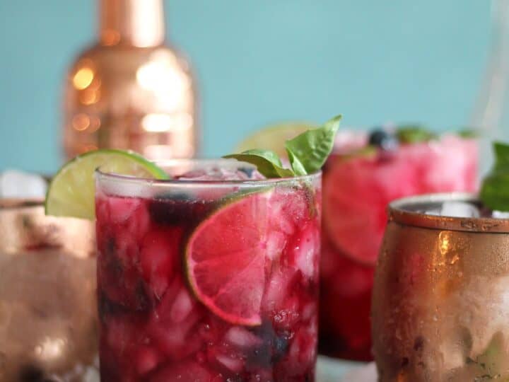 A glass of berry cocktail with lime slices and basil leaves, surrounded by ice cubes. Copper mugs and a cocktail shaker are in the background.