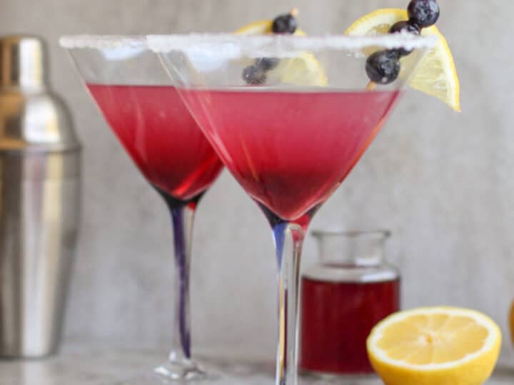 Two martini glasses with red cocktails, garnished with lemon slices and blueberries, sit on a marble surface. A cocktail shaker, lemon half, and small jar are in the background.