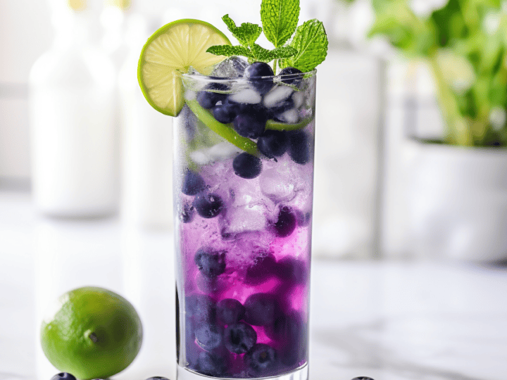 A tall glass with a blueberry drink garnished with lime, mint, and ice. Fresh blueberries and a lime are scattered on the marble surface. Blurred plants in the background.