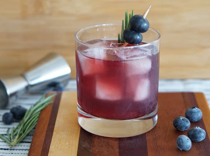A glass of iced purple cocktail garnished with blueberries and rosemary on a wooden board, with a jigger and blueberries in the background.