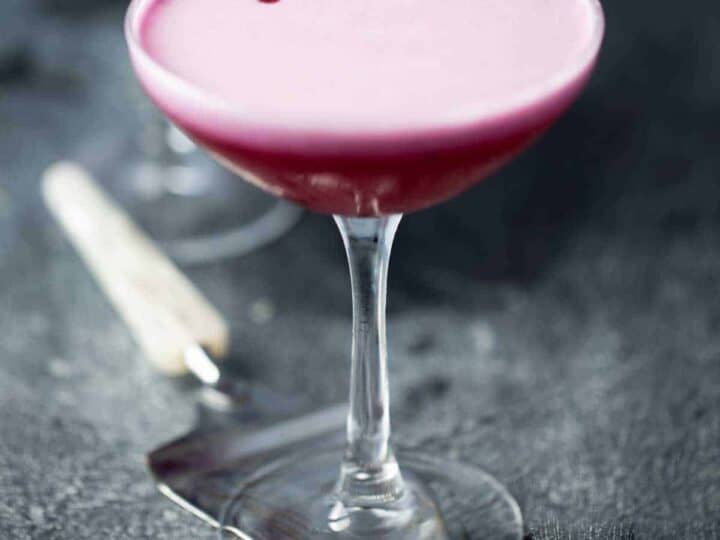 A coupe glass filled with a pink foamy cocktail, placed on a gray surface, with a metal bar tool next to it.