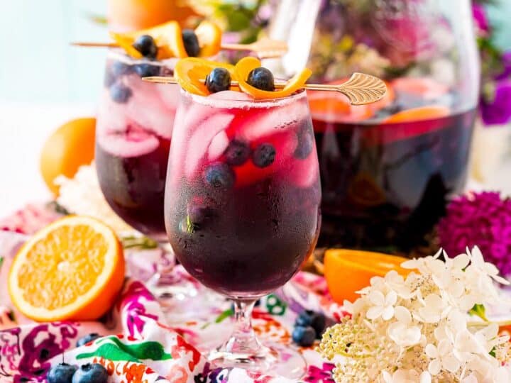 Two glasses of berry sangria with ice and fruit garnishes sit on a floral tablecloth near halved orange and flowers.