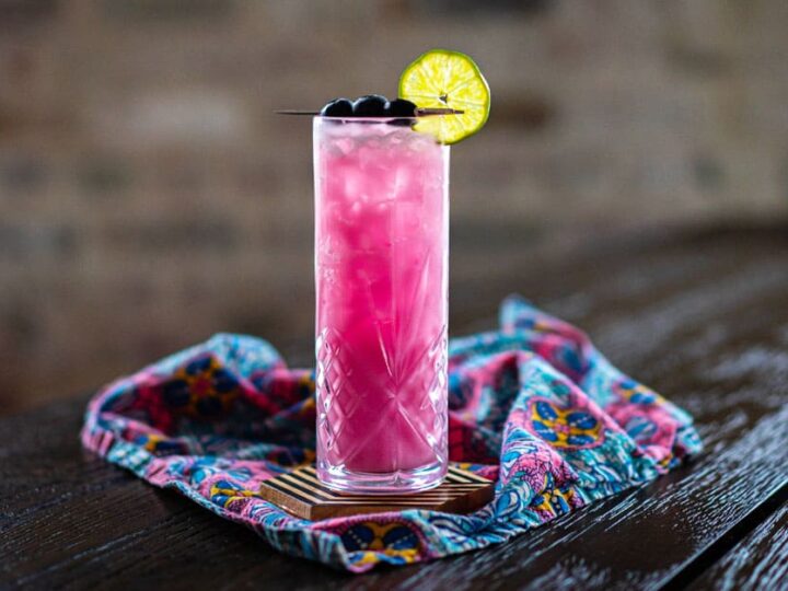 A tall glass of pink iced drink garnished with a lime wheel and blueberries, placed on a patterned cloth on a dark wooden table.