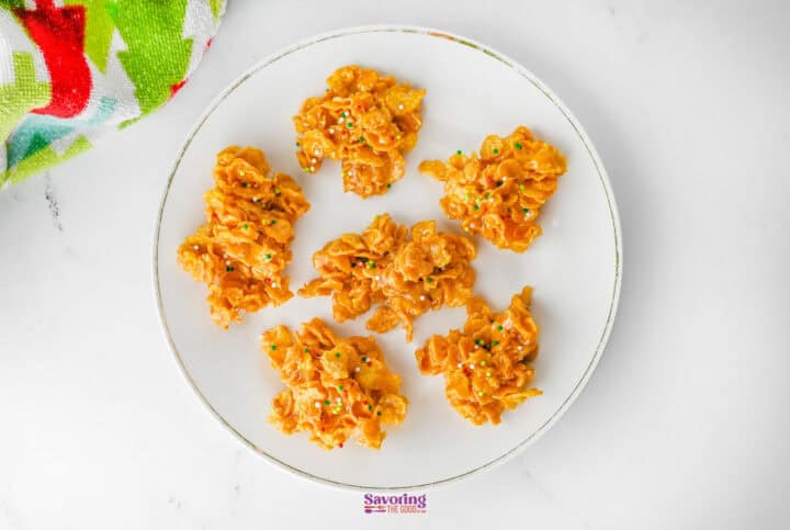 A plate with six clusters of orange-colored crispy treats, likely made from cornflake candy, rests on a white surface. A colorful napkin is partially visible in the corner, adding a splash of whimsy to the scene.