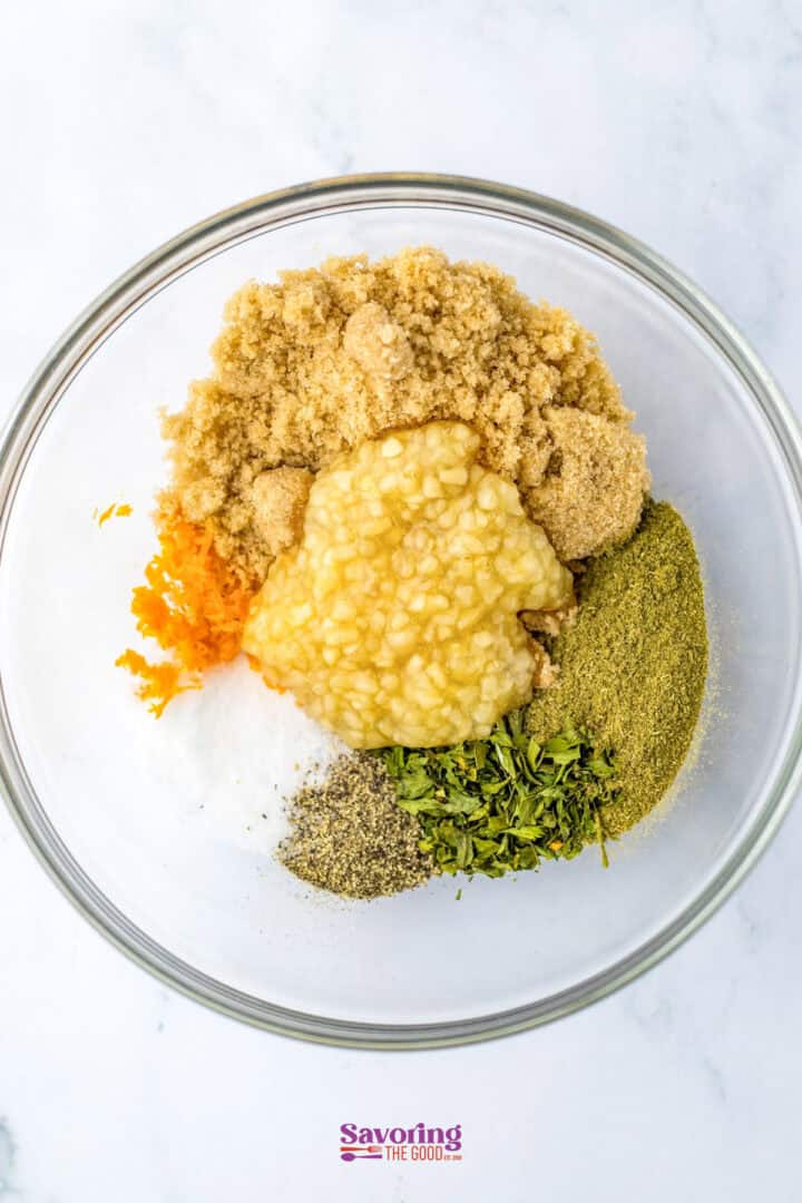 A glass bowl with brown sugar, orange zest, minced garlic, pepper, parsley, and herbs-perfect for seasoning your Christmas chicken. Ingredients are neatly arranged in separate piles on a white surface.