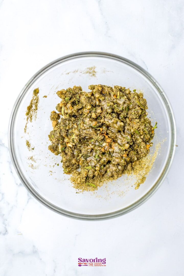 A clear glass bowl filled with a textured green mixture, possibly herbs and spices for seasoning Christmas chicken, rests on a white surface.