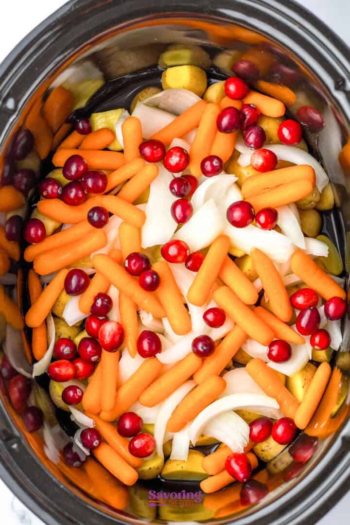 A slow cooker containing baby carrots, cranberries, and onion slices creates a festive medley reminiscent of a Christmas chicken feast.