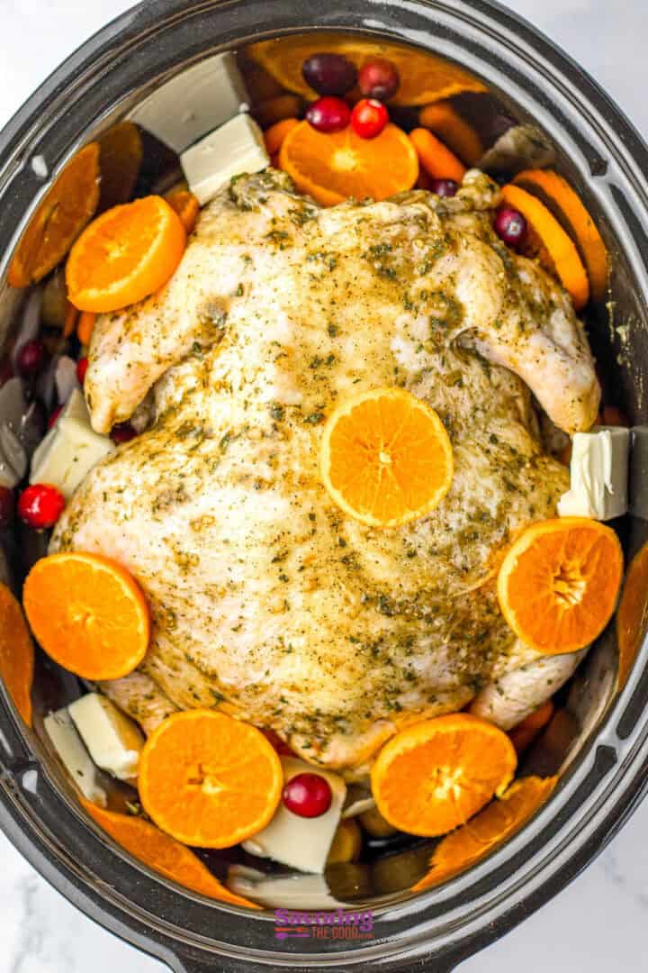 Whole chicken seasoned with herbs, surrounded by orange slices, cranberries, and butter in a slow cooker.
