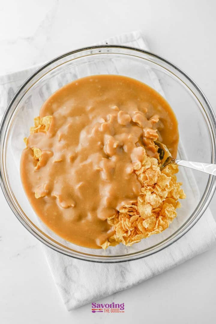 A glass bowl cradles a partially mixed cornflake candy creation, where a luscious peanut butter mixture hugs the crunchy flakes. Resting on a white cloth, the spoon stands ready to combine these delightful ingredients into an irresistible treat.