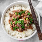 A bowl of slow cooker Mongolian beef atop rice, garnished with chopped green onions and sesame seeds, with chopsticks elegantly placed to the side.