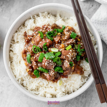 A bowl of slow cooker Mongolian beef atop rice, garnished with chopped green onions and sesame seeds, with chopsticks elegantly placed to the side.