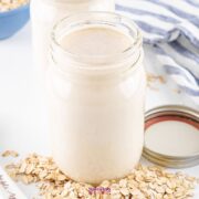 A glass jar filled with creamy oatmeal milk is surrounded by scattered oats and a jar lid on a white surface. A striped cloth adds a touch of texture in the background.